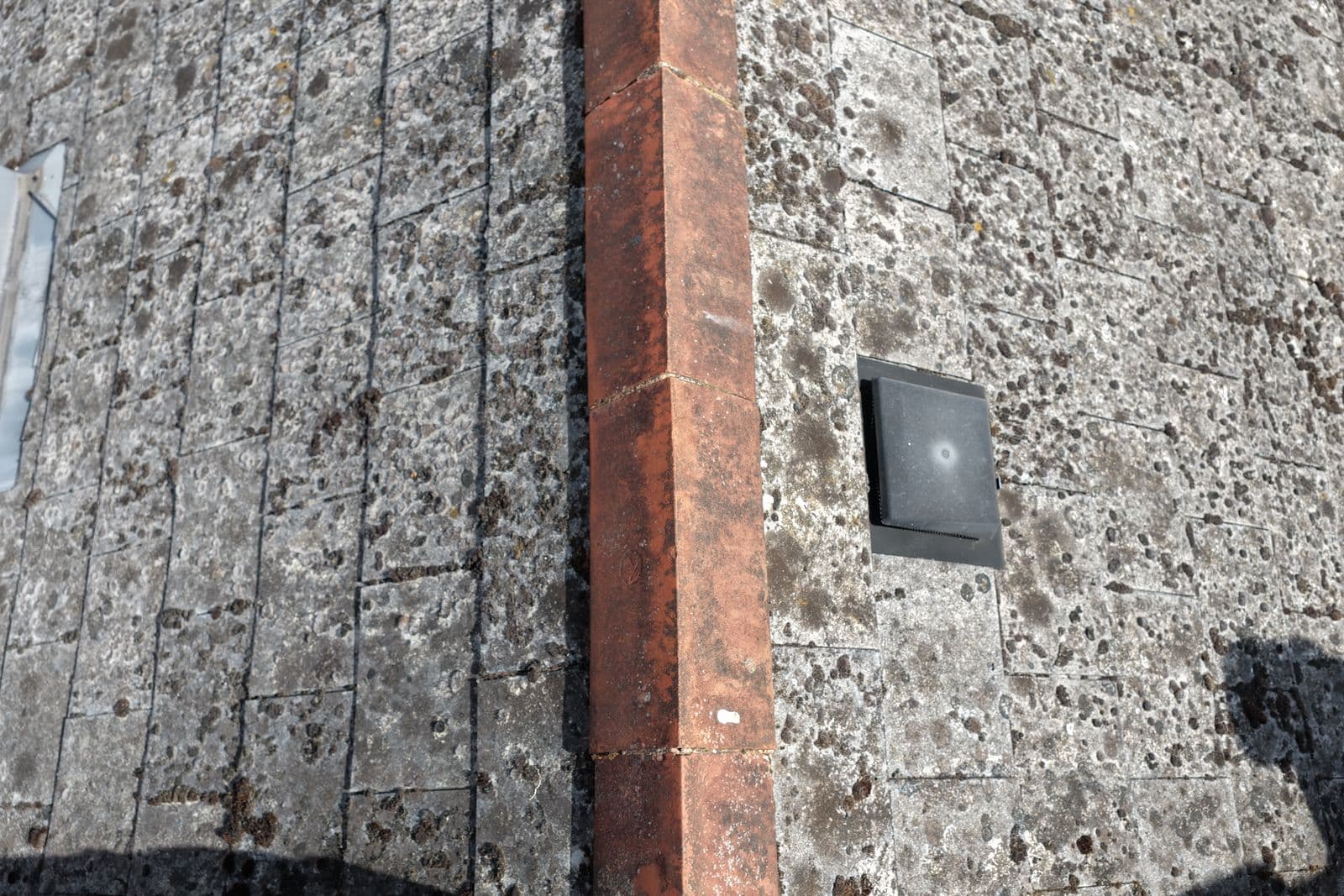 Close aerial detail of a ridge line running across a lichen covered tiled roof slope, with a roof vent alongside