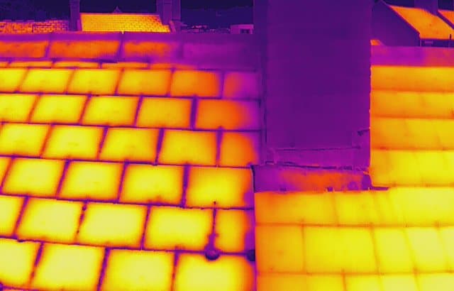 Radiometric thermal image of a slate roof slope beside a chimney stack, the slates rendered in bright yellow and the cooler masonry of the stack in purple