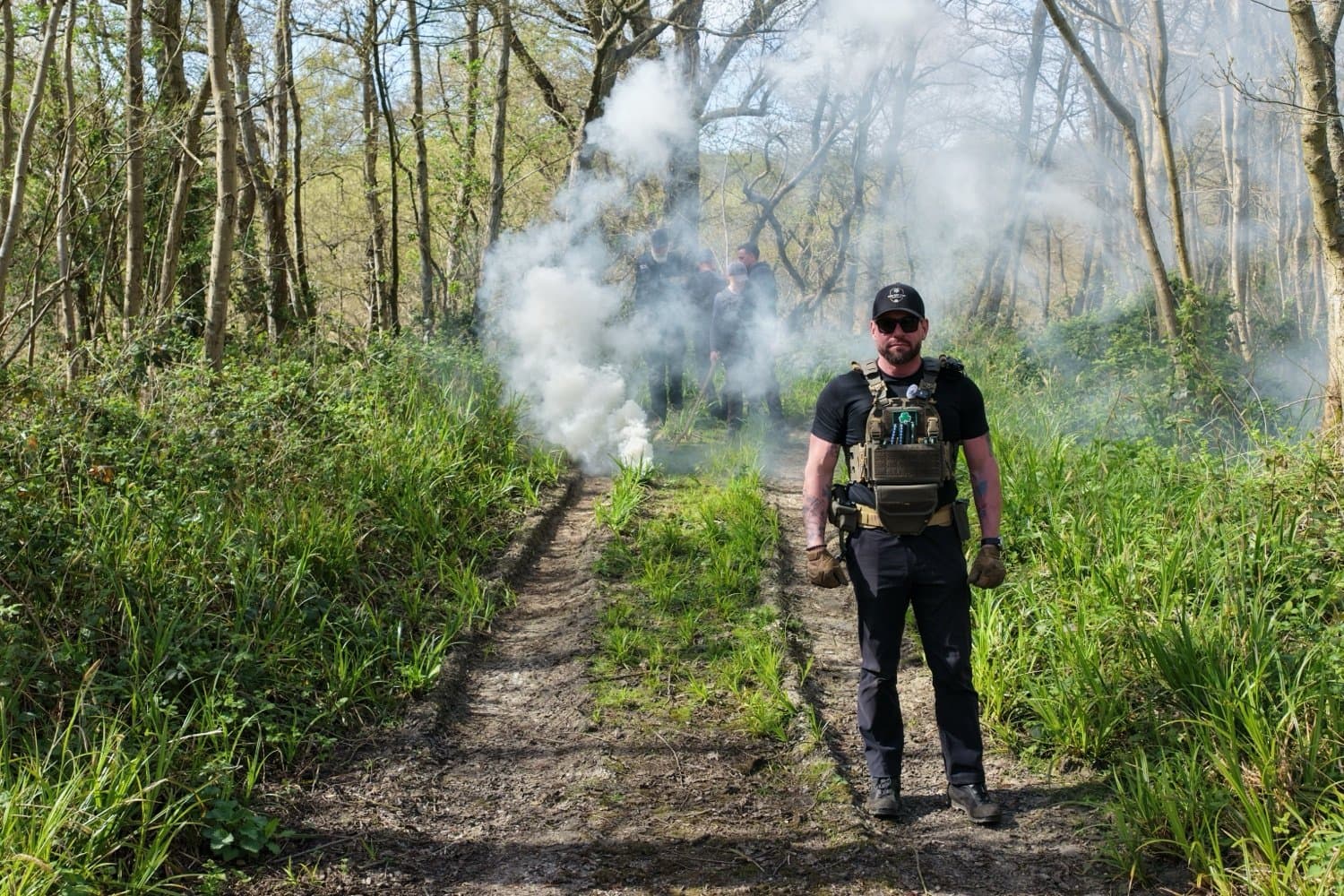An operator in a plate carrier and cap stands on a woodland track at the training area, with a white smoke plume rising behind him and the course group beyond