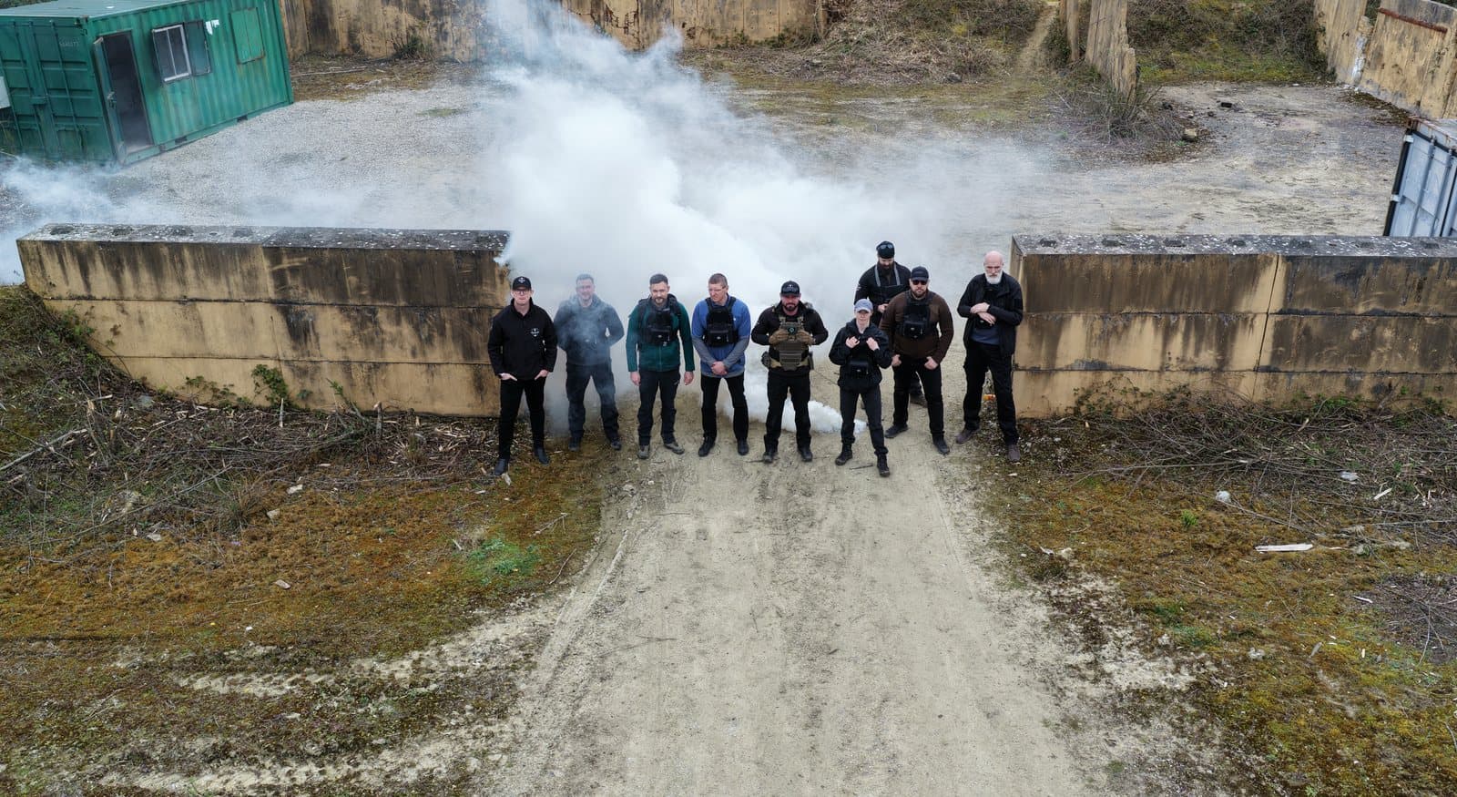 Aerial view looking down a track at a military training area, with a course group formed up along a concrete revetment and a white smoke plume rising beside them